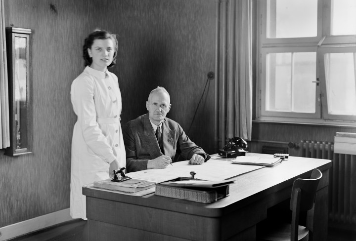 Schwarzweiß-Bildes eines am Schreibtisch sitzenden Mannes und einer daneben stehenden Frau in einem Büro