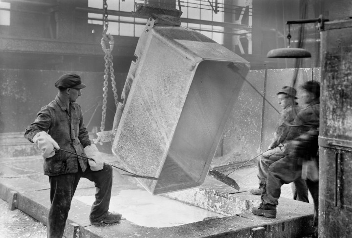 Schwarzweiß-Foto von Männern in einer Fabrik, die einen Grubenwagen mit glühendem Inhalt auskippen