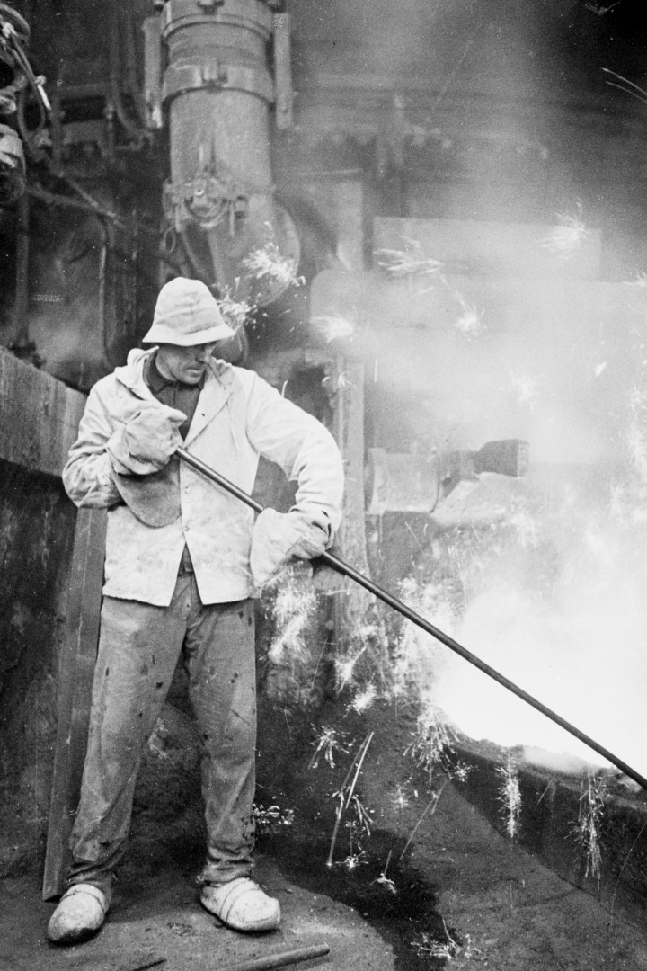 Schwarzweiß-Bild eines Arbeiters, der eine Stange in ein Feuer hält