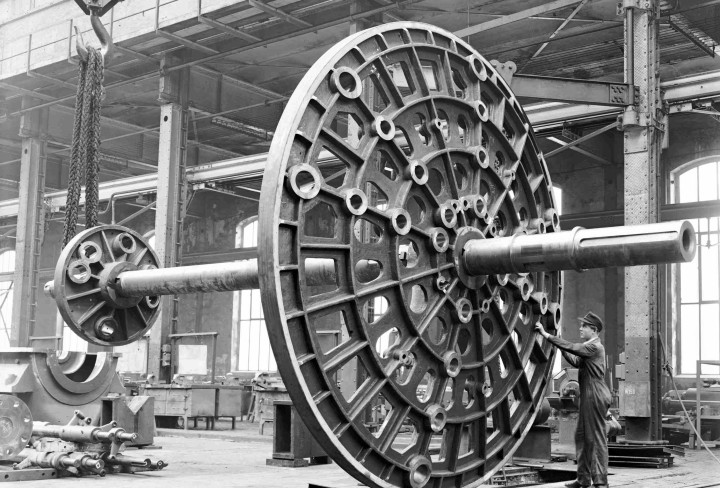 Schwarzweiß-Foto eines großen Maschinenteils, daneben steht ein Arbeiter