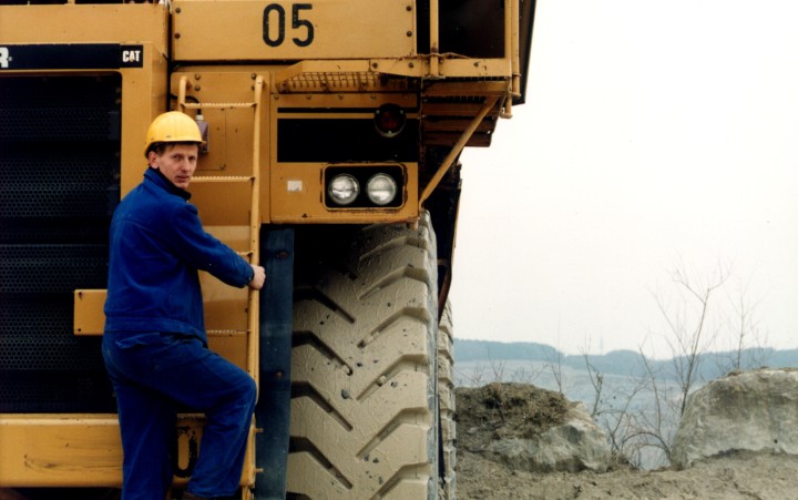 Farbfotografie eines Arbeiters mit Blaumann und Helm auf einem Industriefahrzeug
