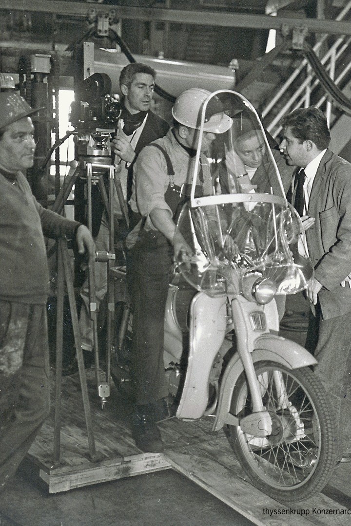 Schwarzweiss-Foto zeigt ein Mann auf einem Moped, andere stehen daneben